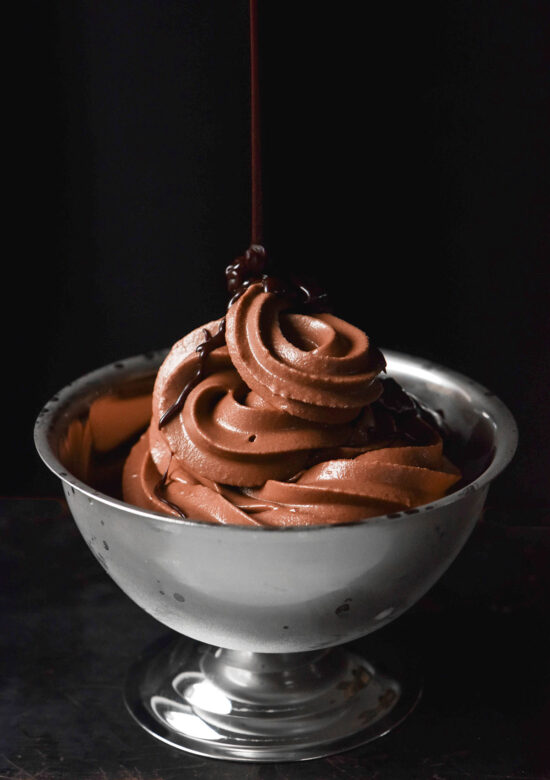 A side on moody image of a frosted silver dessert coupe filled with Ninja Swirl chocolate soft serve. A drizzle of brown butter hot fudge sauce swirls down from the top of the image