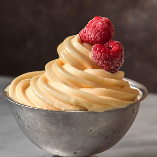 A side on image of a silver dessert coupe filled with Ninja Swirl custard soft serve. The coupe sits on a white marble table against a brown backdrop. Two frozen raspberries sit atop the frozen custard.