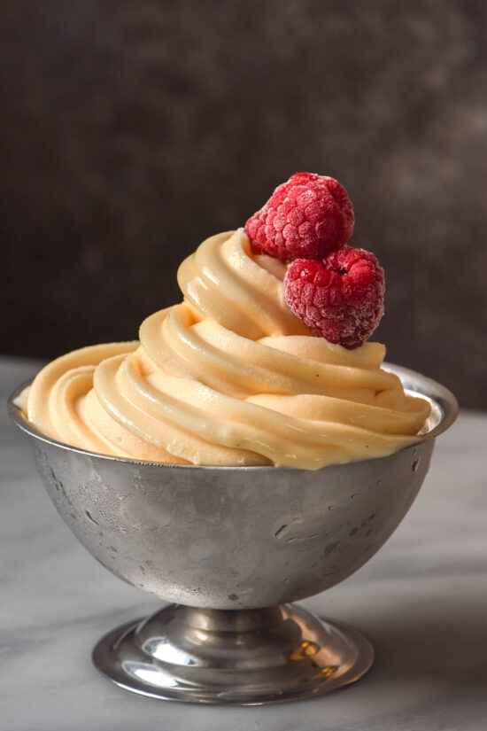A side on image of a silver dessert coupe filled with Ninja Swirl custard soft serve. The coupe sits on a white marble table against a brown backdrop. Two frozen raspberries sit atop the frozen custard.