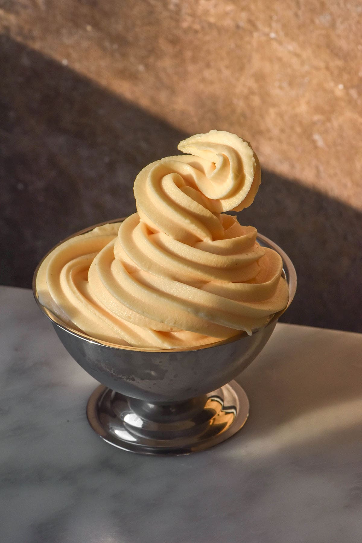 A side on image of a silver dessert coupe filled with swirls of Ninja Swirl frozen custard. The coupe sits on a white marble table against a brown backdrop in contrasting sunlight.