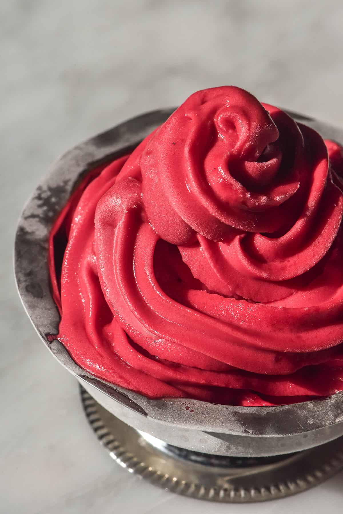 A side on close up image of raspberry sorbet soft serve in a silver dessert coupe on a white marble table