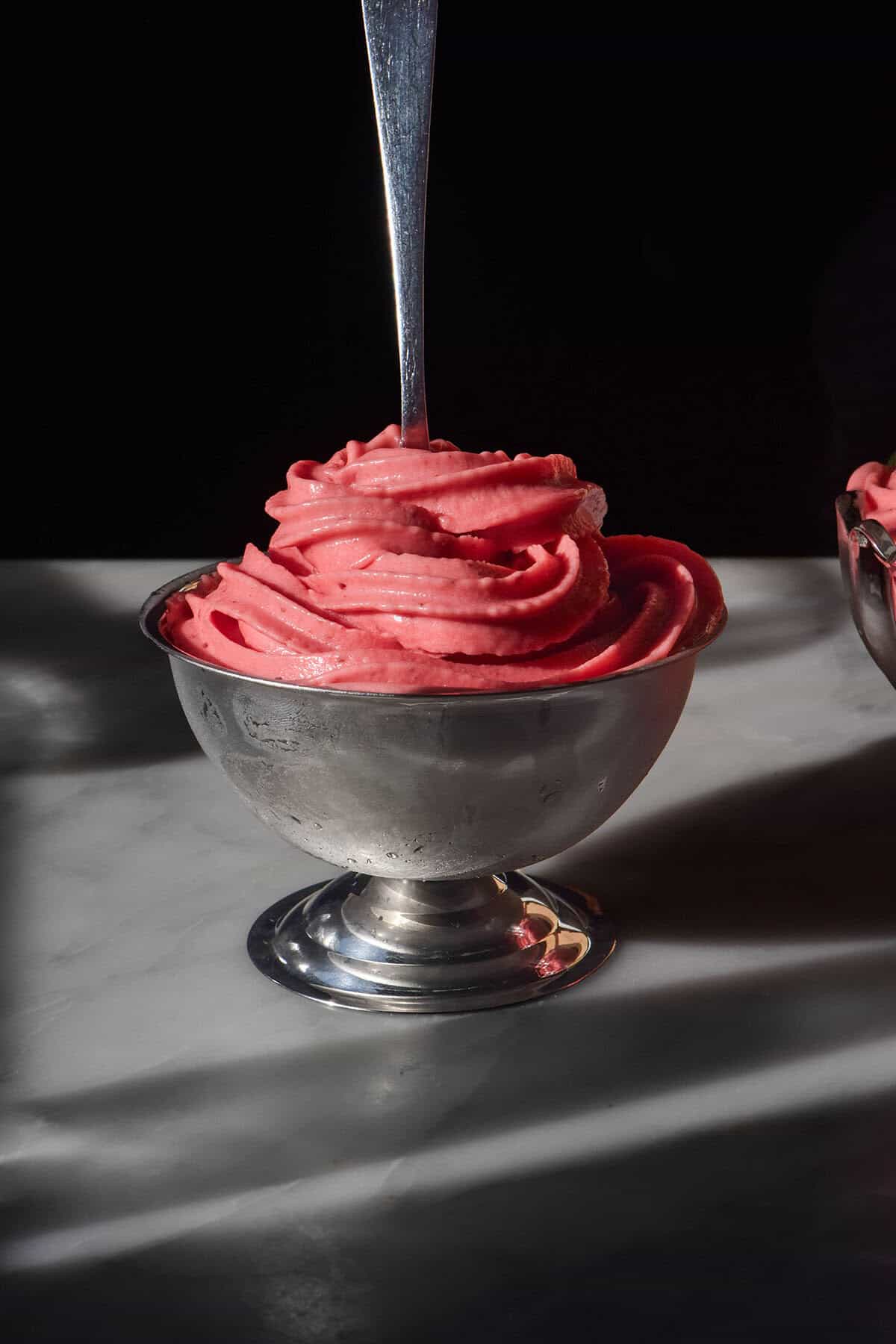 A side on image of a silver dessert coupe on a white marble table against a black backdrop. The coupe is filled with strawberry soft serve ice cream and a spoon sits in the centre of the soft serve.