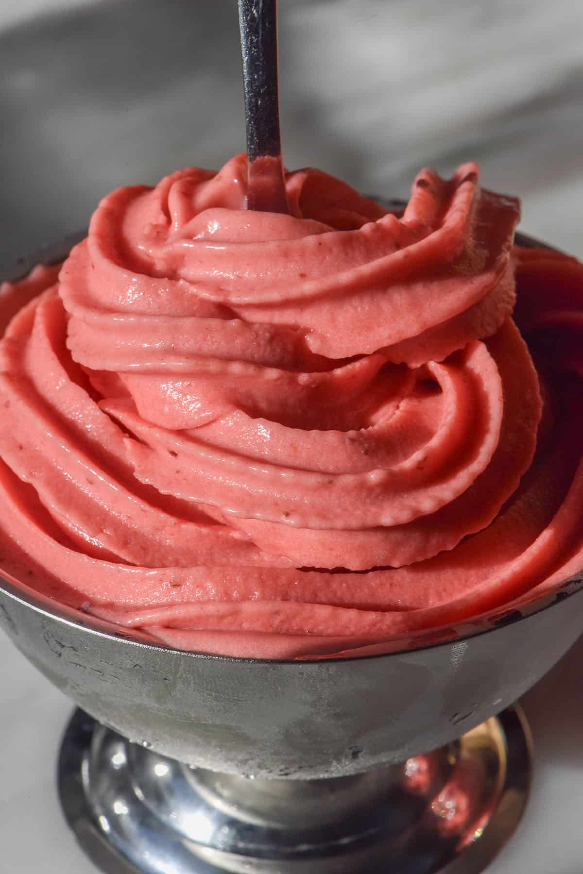 An image of strawberry soft serve ice cream in a silver dessert coupe on a white marble table. A spoon sticks out of the centre of the coupe