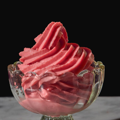 A side on image of a Ninja Swirl strawberry soft serve in a fluted glass dessert cup on a white marble table against a dark backdrop
