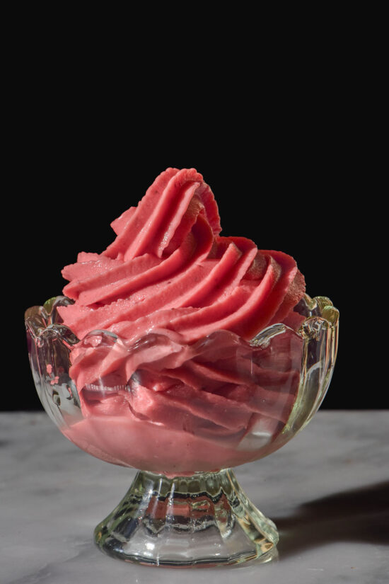 A side on image of a Ninja Swirl strawberry soft serve in a fluted glass dessert cup on a white marble table against a dark backdrop