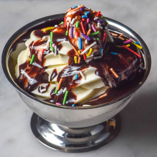 A side on image of a silver dessert coupe on a white marble table. The dessert coupe is filled with Ninja Swirl vanilla soft serve and topped with hot fudge sauce and rainbow sprinkles