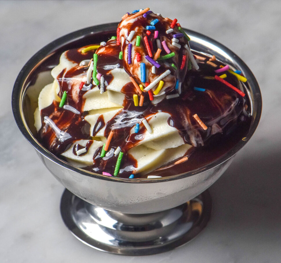 A close up image of Ninja Swirl vanilla soft serve in a silver dessert coupe on a white marble table. The ice cream is topped with hot fudge sauce and rainbow sprinkles