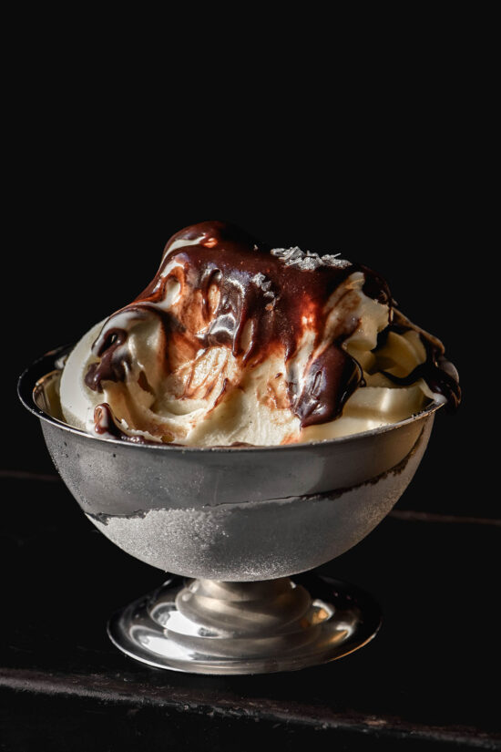 A side on image of a dessert coupe filled with Ninja Swirl vanilla soft serve topped with a hot fudge sauce. The coupe sits against a dark backdrop on a dark table