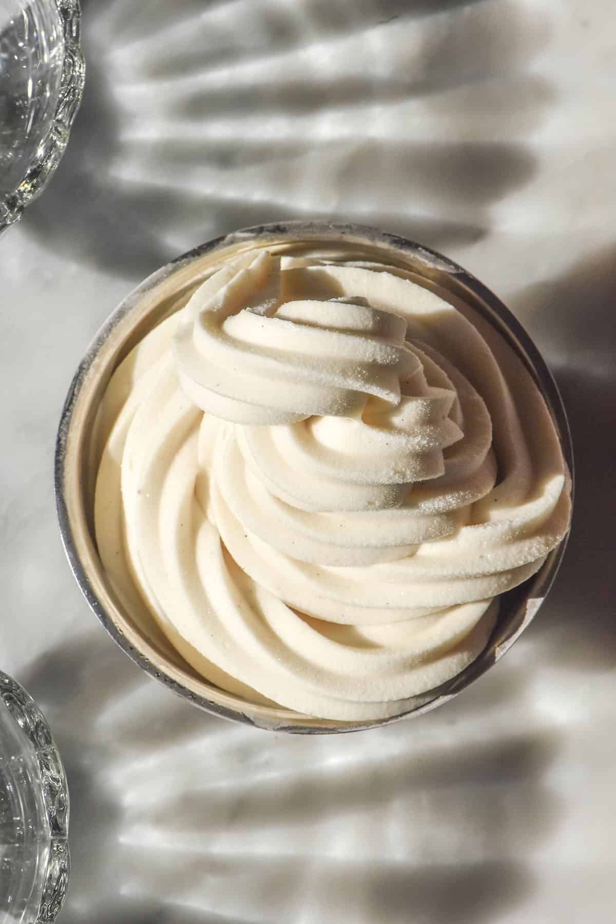 An aerial image of a silver dessert coupe filled with a vegan Ninja Swirl vanilla soft serve. Two scalloped glasses sit to the left of the coupe on either side, creating scalloped light and shadow patterns on the white marble table
