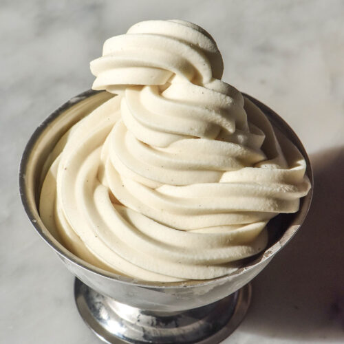 A brightly lit side on image of a silver dessert coupe filled with a vegan soft serve ice cream. The coupe sits in the centre of a white marble table.