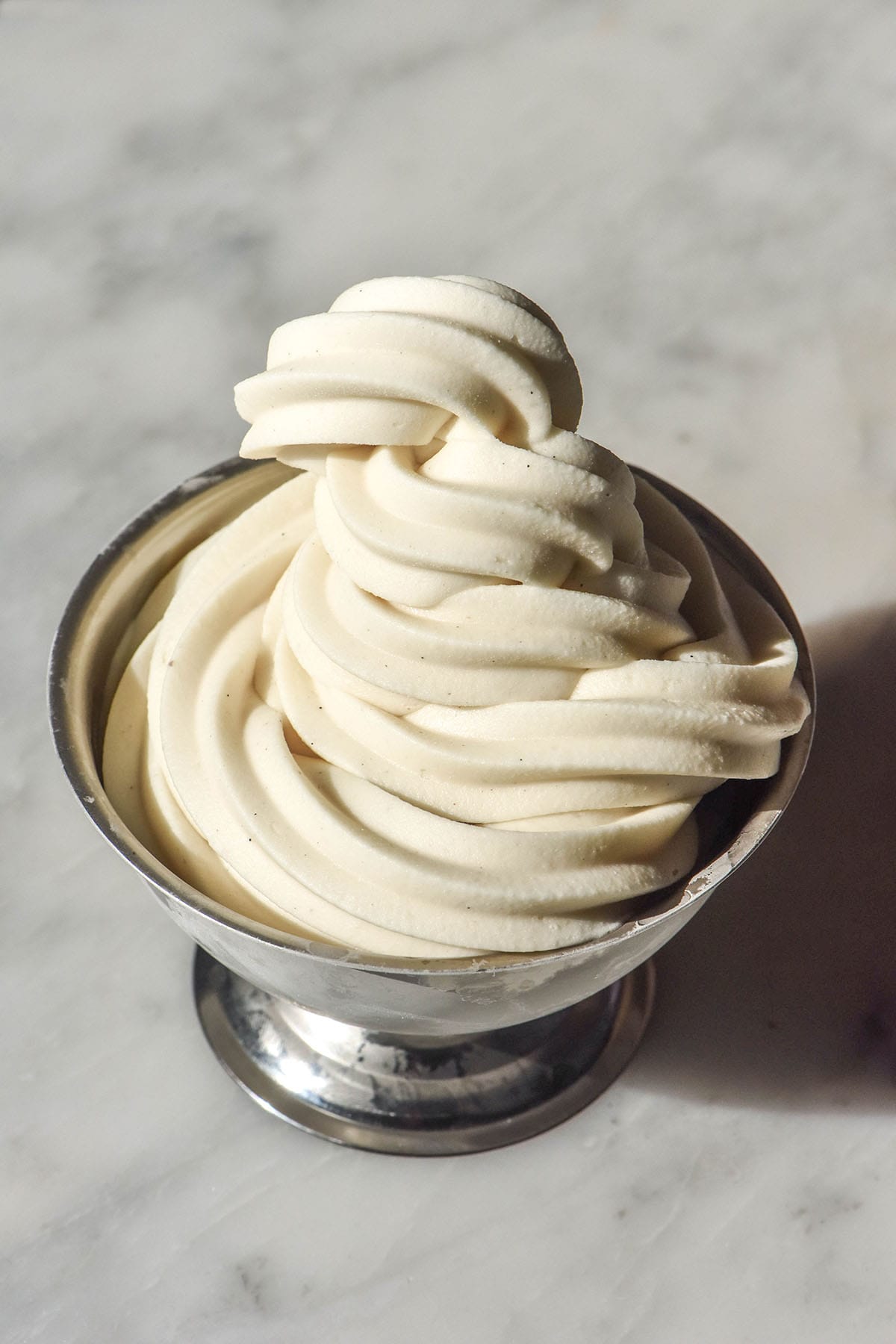 A brightly lit side on image of a silver dessert coupe filled with a vegan soft serve ice cream. The coupe sits in the centre of a white marble table.