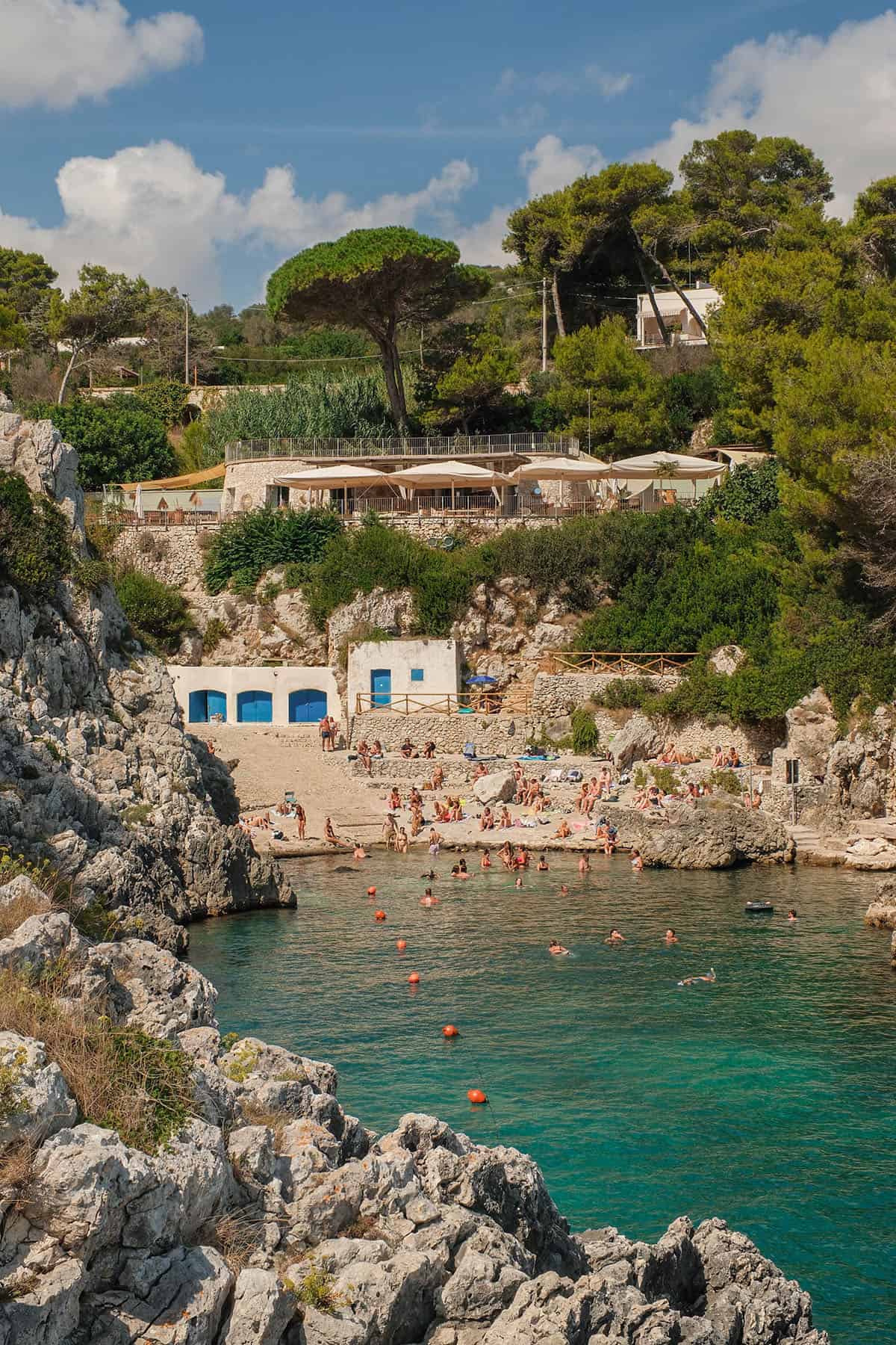 An image looking back at Cala dell'Acquaviva from the rocky coastal outcrop on the other side of the cove. The sky is blue and the water is filled with people.