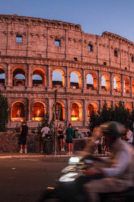 An image of a man on a vespa riding past the Colosseum at sunset in Rome, Italy