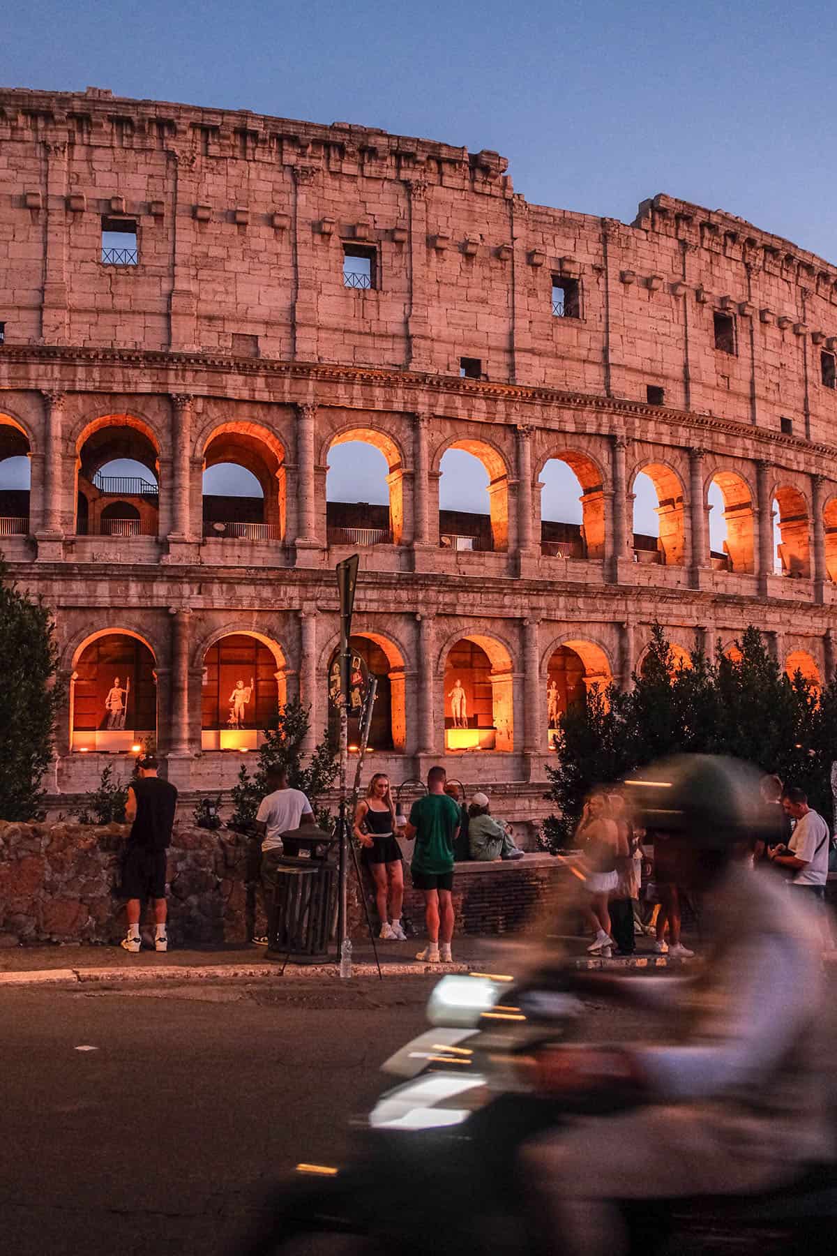 An image of a man on a vespa riding past the Colosseum at sunset in Rome, Italy