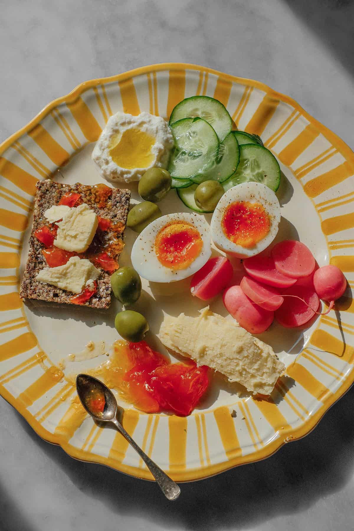 An aerial image of a yellow and white striped snack plate with jammy eggs, cheese, pickles, marmalade, olives and high protein crackers.
