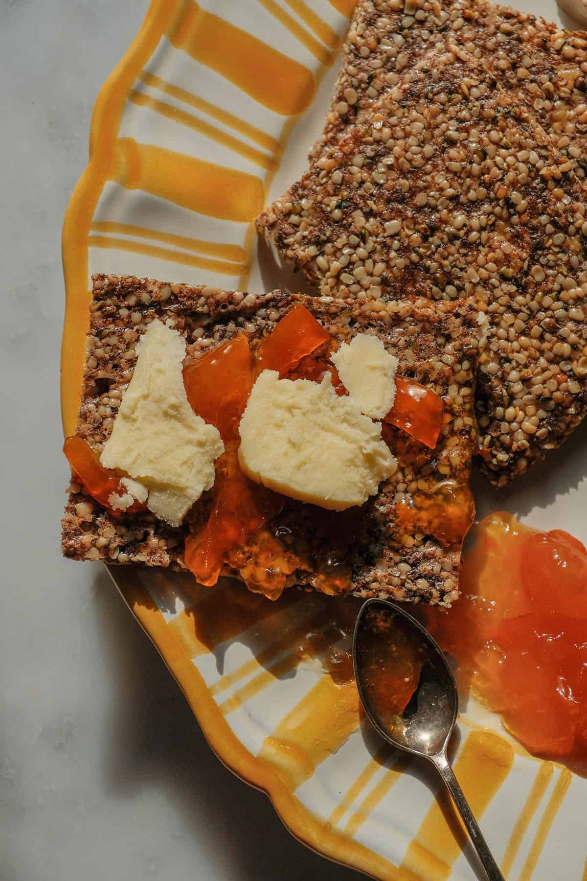 An aerial image of high protein crackers on a yellow and white striped plate. The crackers are topped with cheese and cumquat marmalade and the plate sits atop a white marble table.
