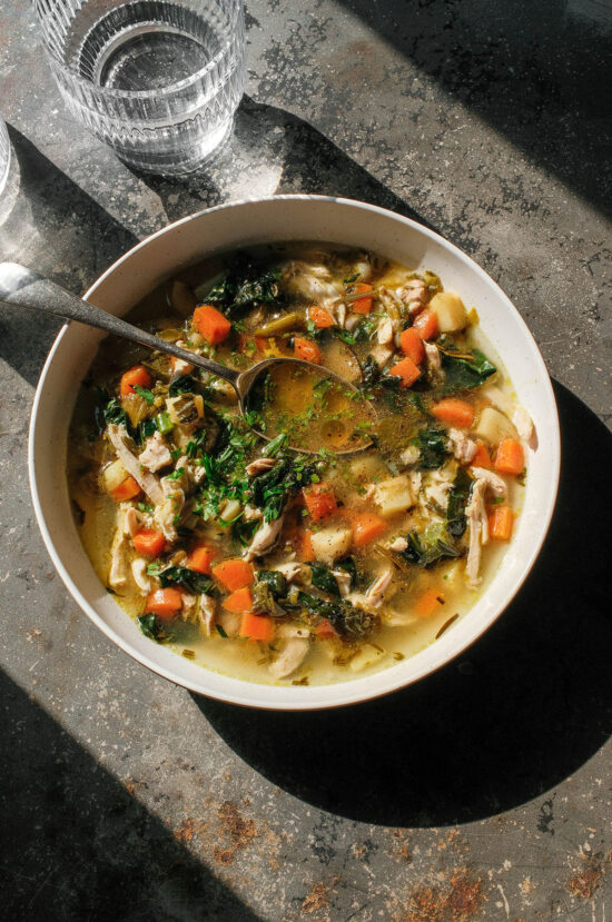 An aerial image of a white bowl of low FODMAP chicken soup on a dark grey steel backdrop in contrasting sunlight.