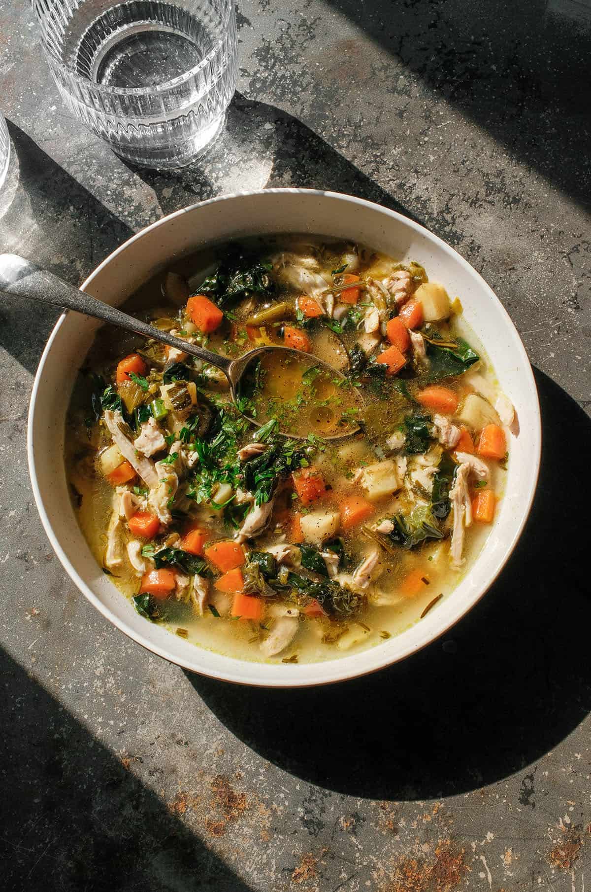 An aerial image of a white bowl of low FODMAP chicken soup on a dark grey steel backdrop in contrasting sunlight.