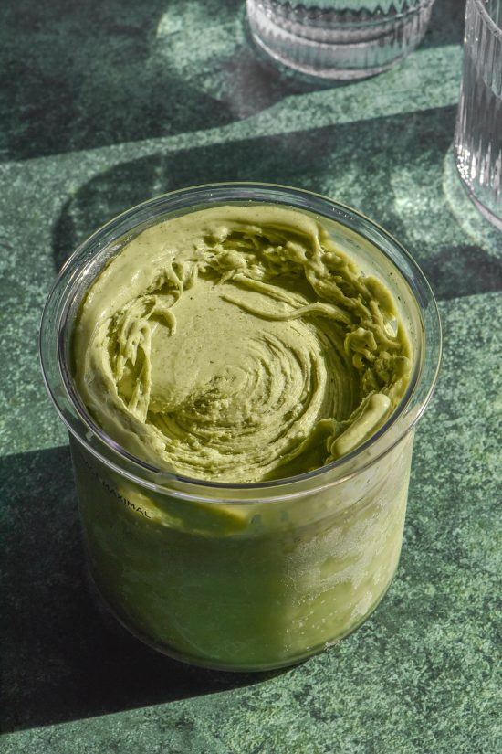 A brightly lit aerial image of a tub of high protein matcha ninja Creami ice cream on a dark green backdrop. Two sunlit glasses of water sit to the right of the image