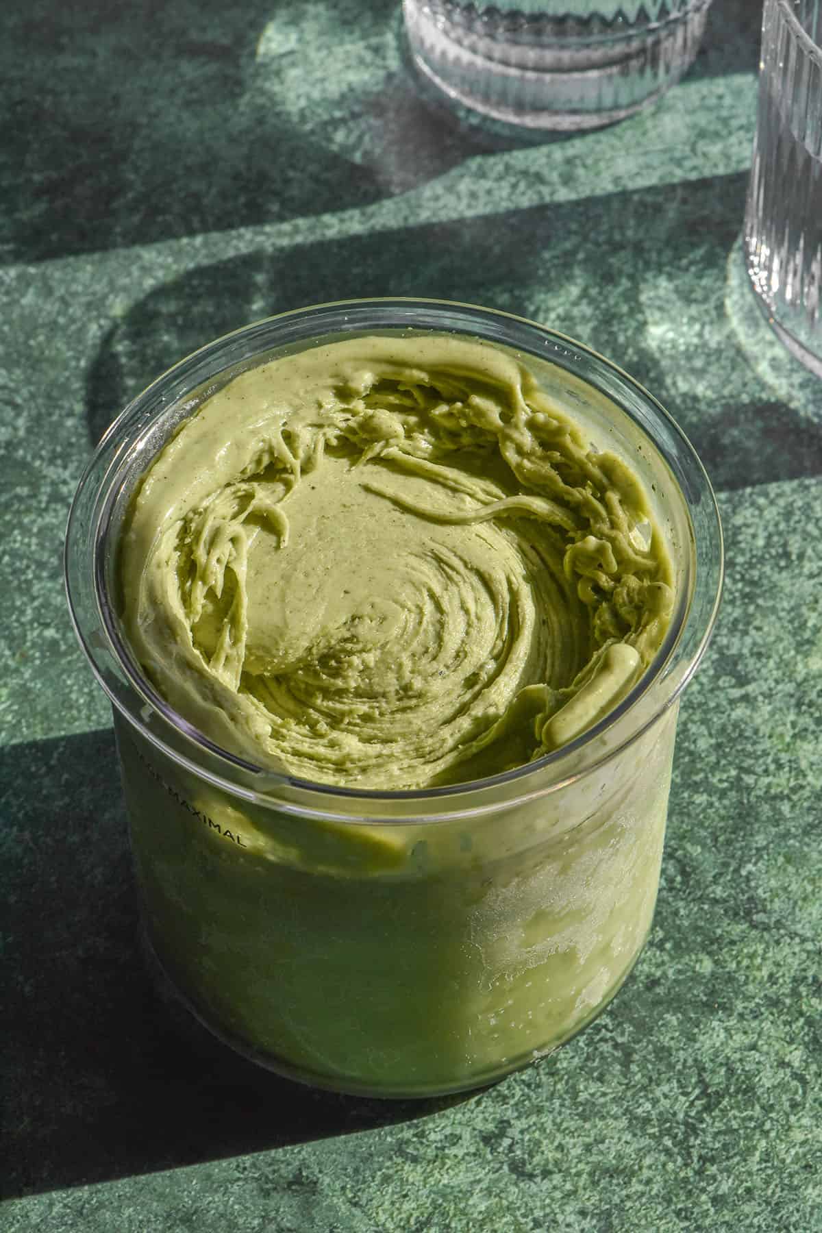 A brightly lit aerial image of a tub of high protein matcha ninja Creami ice cream on a dark green backdrop. Two sunlit glasses of water sit to the right of the image