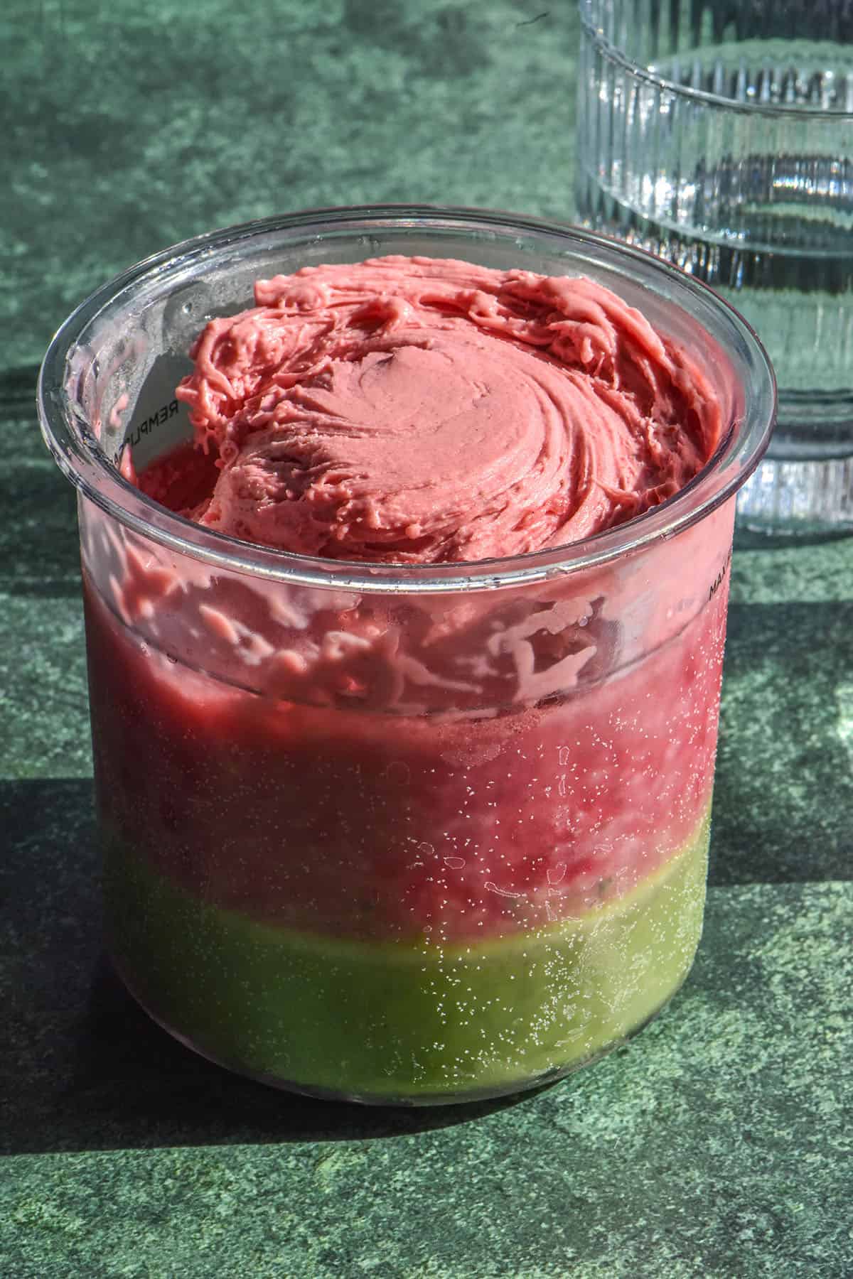 A side on image of a strawberry matcha Ninja Creami on an olive green backdrop. A sunlit glass of water sits in the back right of the image