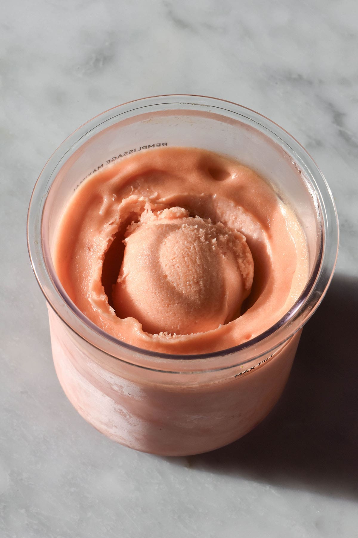 An aerial image of a tub of peach Ninja Creami ice cream atop a sunlit white marble table