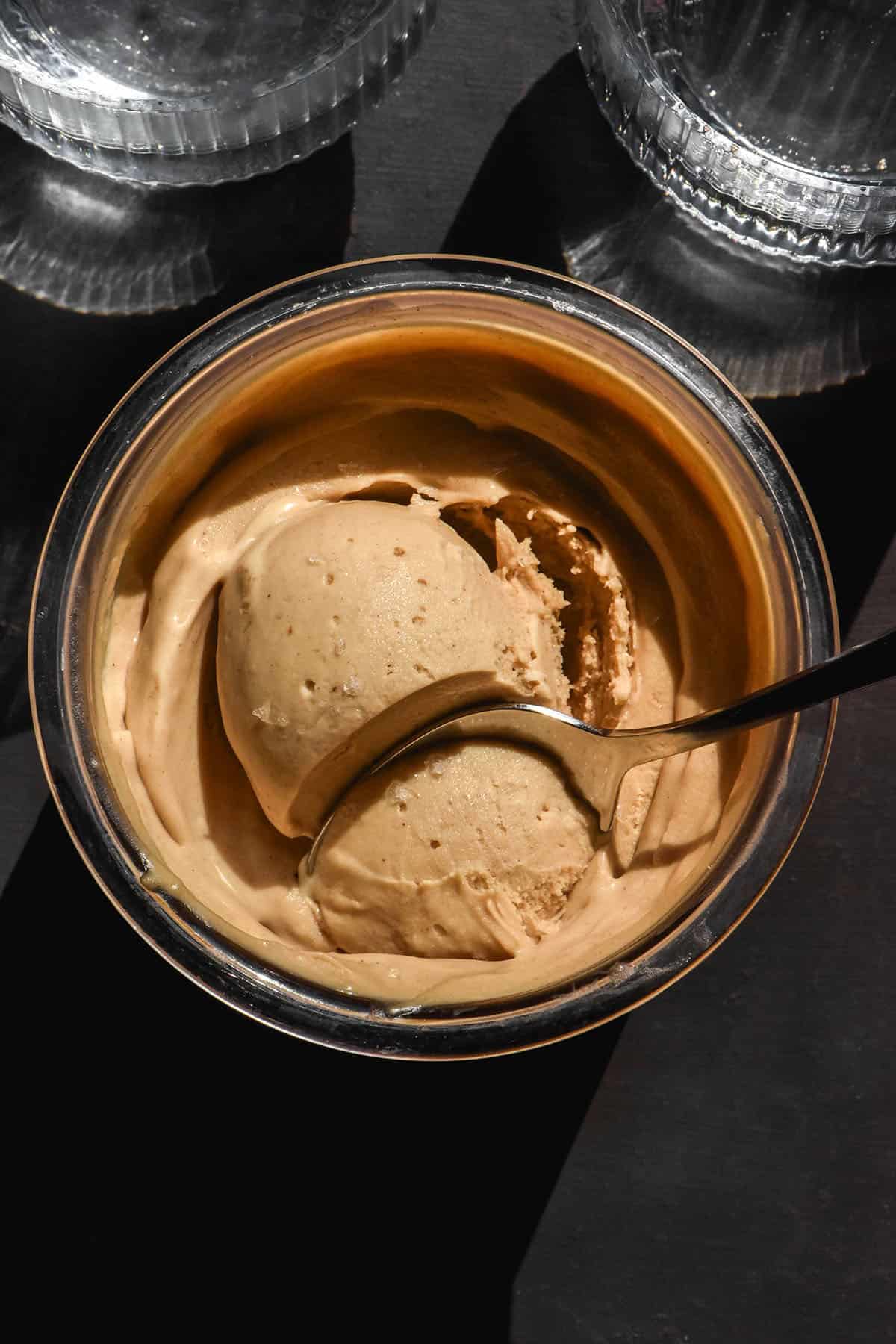 An aerial image of a tub of brown butter Ninja Creami ice cream on a black backdrop with two sunlit glasses of water in the top of the image