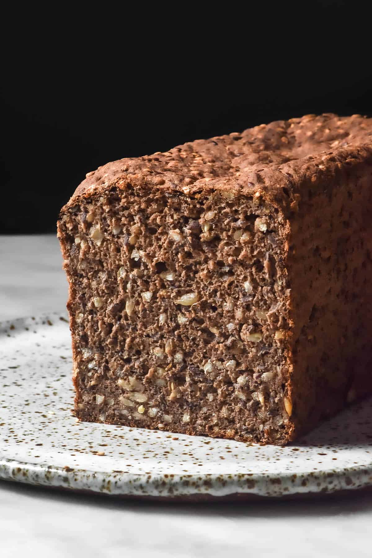 A side on image of a loaf of gluten free Danish style sourdough seeded 'rye' on a white marble table against a dark backdrop