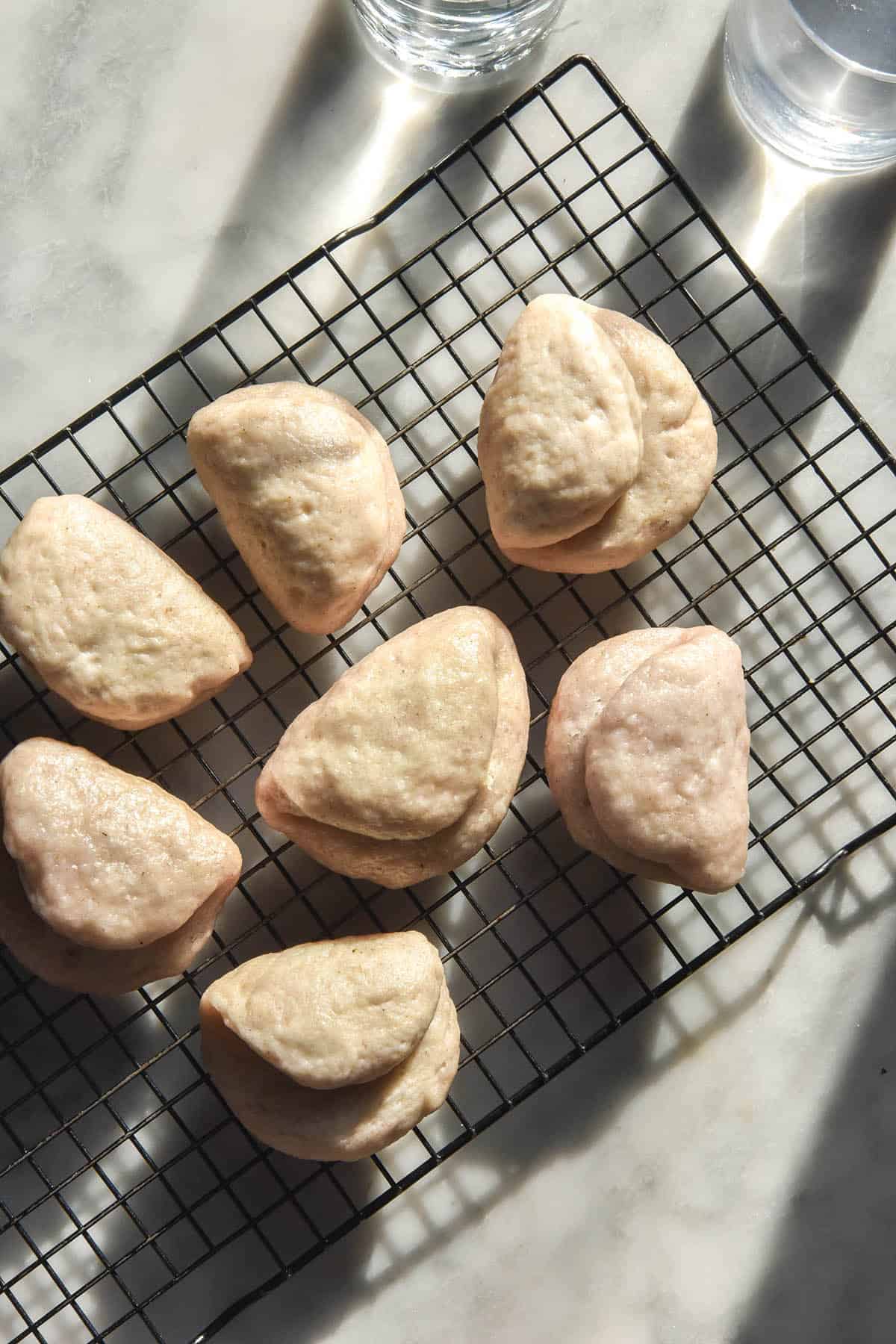 An aerial image of gluten free sourdough bao buns in bright contrasting sunlight. They sit on a wire cooling rack atop a white marble table and two sunlit water glasses sit in the top of the image.