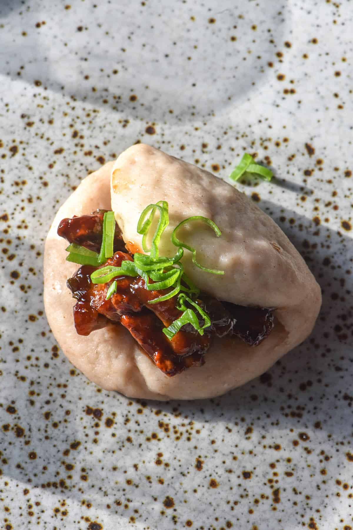 An aerial image of a gluten free sourdough bao bun on a white speckled ceramic plate. The bun is stuffed glazed tofu and spring onion greens.