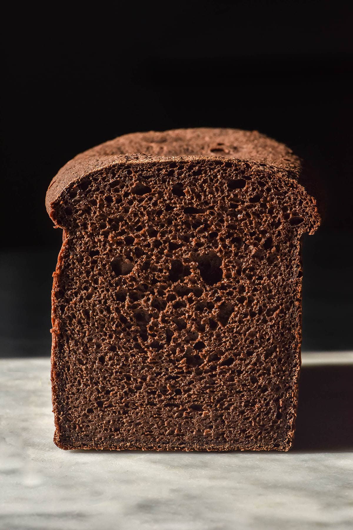 A side on image of a loaf of gluten free sourdough pumpernickel bread on a white marble table against a black backdrop