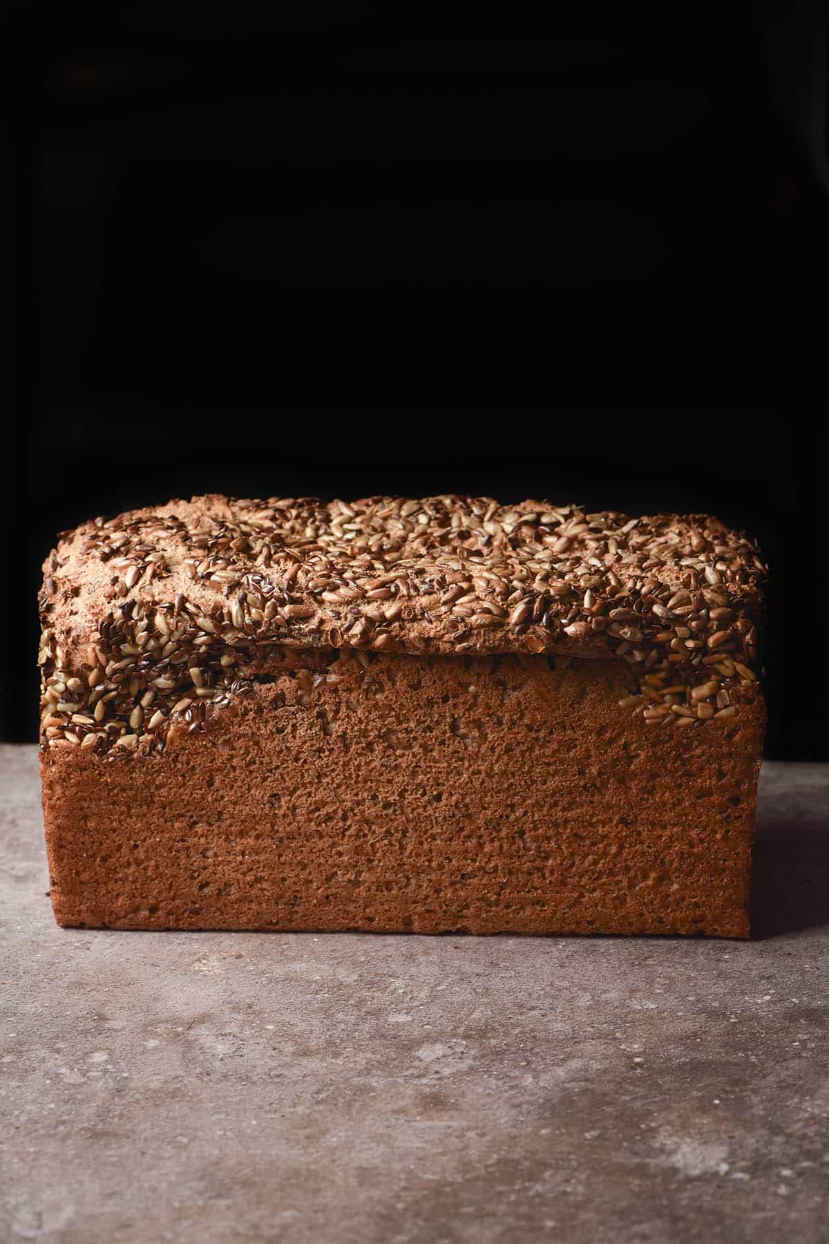 A moody side on image of a gluten free sourdough seeded loaf on a brown table against a black backdrop